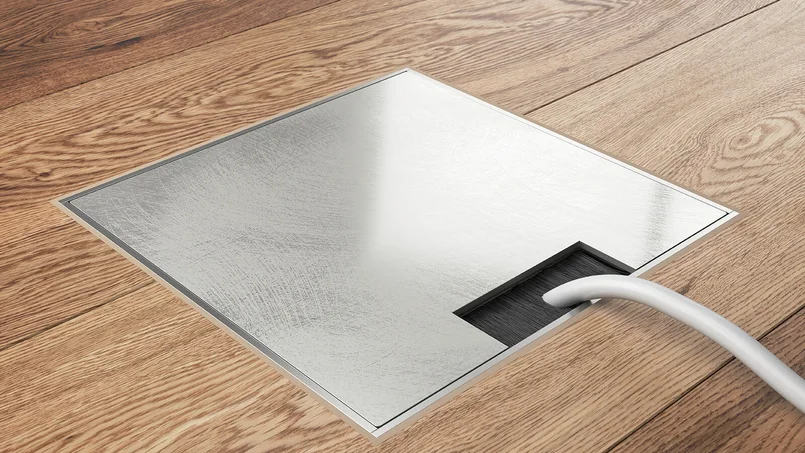 Ein quadratischer Bodeneinsatz mit gebürsteter Metallabdeckung ist in einen Holzfußboden eingelassen; rechts führt ein Kabel durch die offene Aussparung. Das Bild zeigt eine moderne, bündig integrierte Bodensteckdose in Detailansicht.
