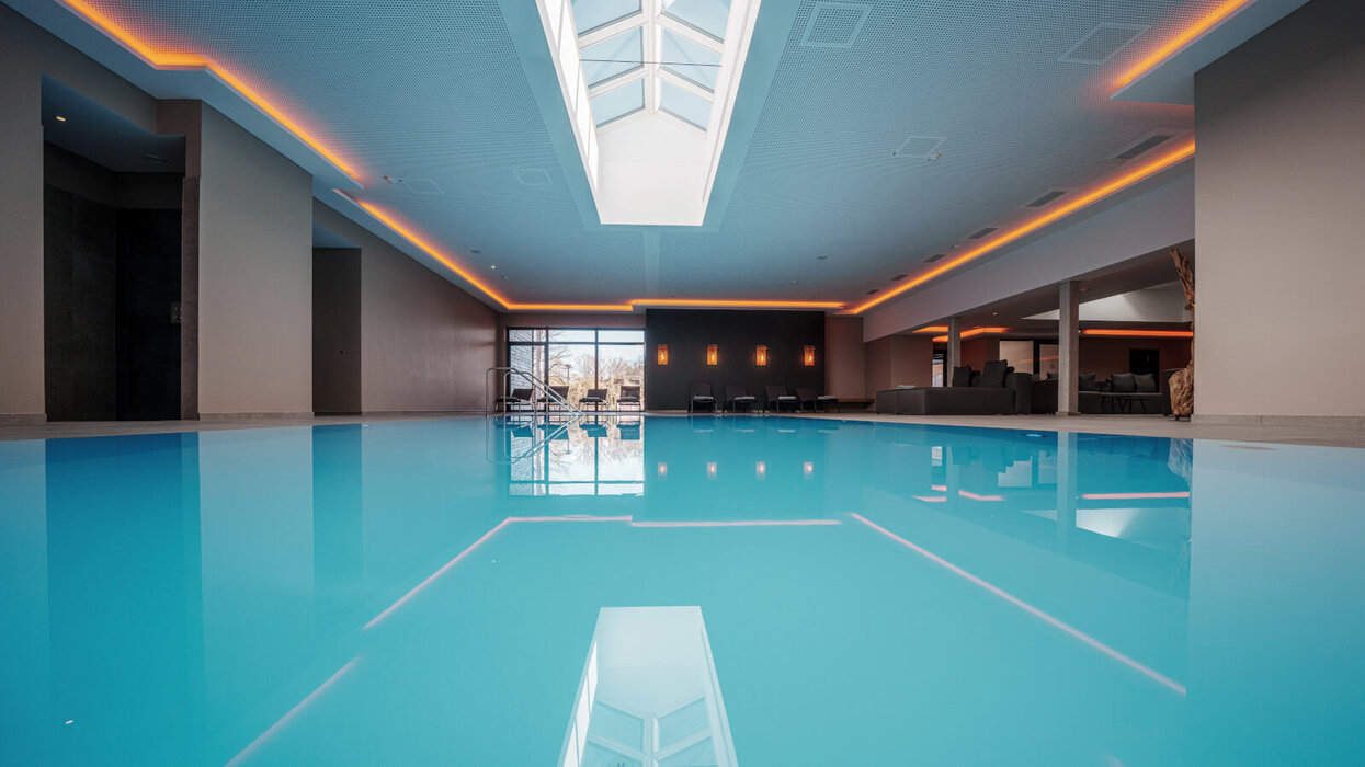 Zu sehen ist ein moderner Indoor-Pool mit großem Wasserbecken und indirekter Deckenbeleuchtung. Der Raum wirkt großzügig, ruhig und hochwertig gestaltet.