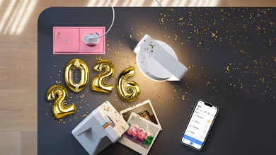 Das Bild zeigt eine Draufsicht auf eine dunkle Tischfläche mit goldenen Luftballons in Form der Zahl „2026“, verstreutem Konfetti, einer rosa Steckdose mit eingestecktem Stecker, einer weißen Leuchte, einer kleinen Fotokamera mit ausgedruckten Fotos sowie einem Smartphone mit geöffneter App.