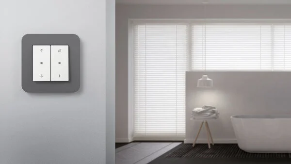 Im Vordergrund des Bildes ist ein graues Gira System 3000 an einer hellgrauen Wand zu sehen. Im Hintergrund des Bildes sieht man eine Badewanne mit einer Sichtschutzwand vor einem großem Fenster. Die Jalousien vor dem Fenster sind geschlossen.