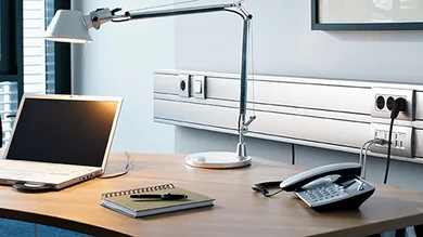 Arbeitsplatz mit Schreibtisch, Laptop, Stehlampe und Telefon. Im Hintergrund ein Leitführungsystem von OBO Bettermann.