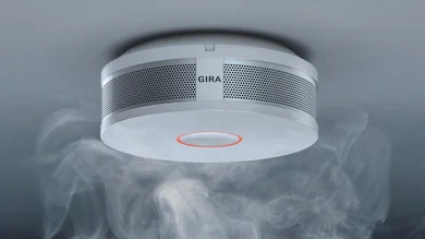 ein weißer Rauchmelder hängt an einer grauen Raumdecke. Rauchschwaden umgeben den Melder.