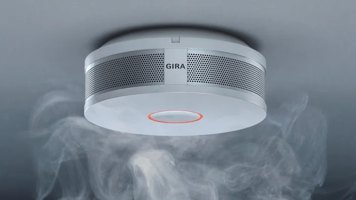 ein weißer Rauchmelder hängt an einer grauen Raumdecke. Rauchschwaden umgeben den Melder.