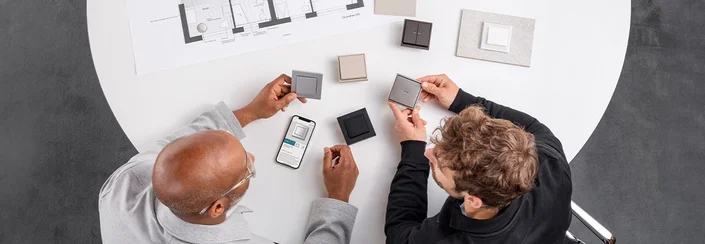 Das Bild zeigt zwei Männer an einem Tisch, die verschiedene Lichtschalter-Designs begutachten. Auf dem Tisch liegen Muster, ein Smartphone mit Produktinformation und ein Bauplan.