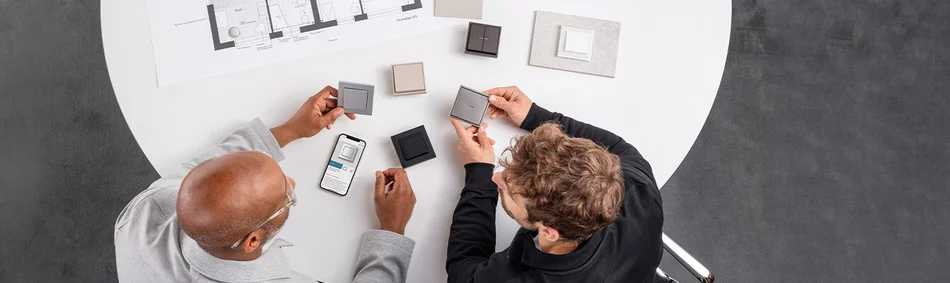 Das Bild zeigt zwei Männer an einem Tisch, die verschiedene Lichtschalter-Designs begutachten. Auf dem Tisch liegen Muster, ein Smartphone mit Produktinformation und ein Bauplan.