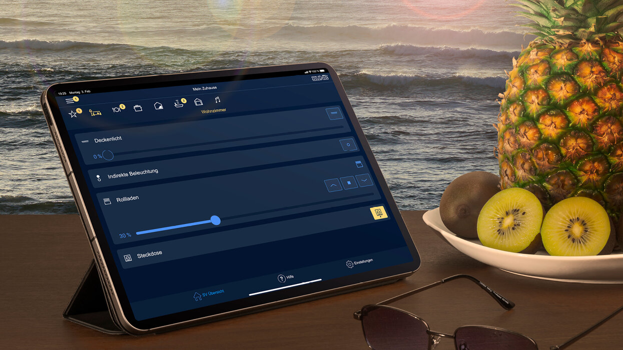 Smart HOME Bundle: JUNG HOME Gateway meets Smart Visu Server Ein Tablet mit geöffneter Smart-Home-Steuerungs-App liegt auf einem Tisch. Im Hintergrund ist ein Meer zu sehen, daneben ein Teller mit Ananas und Kiwis sowie eine Sonnenbrille.