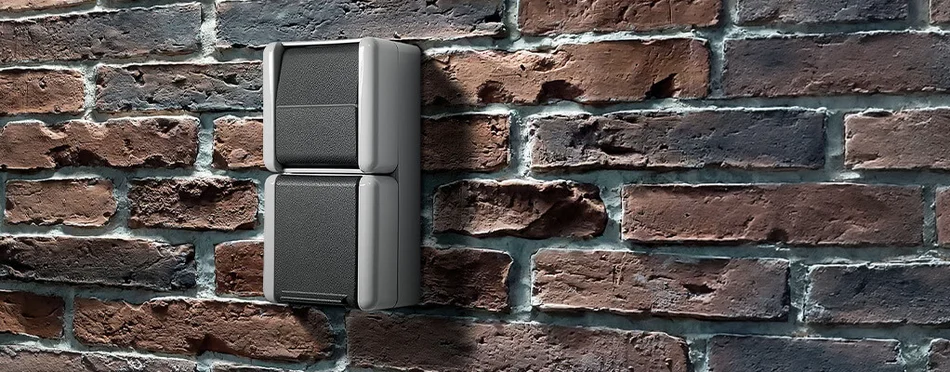 Zu sehen ist ein zwei­teiliger grauer Wandschalter auf einer Backsteinwand. Das Design wirkt robust und schlicht, passend für den Außen- oder Industrieeinsatz.