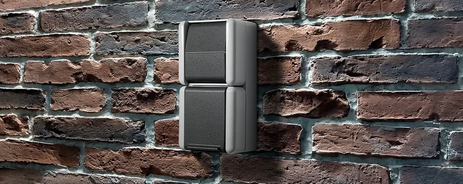 Zu sehen ist ein zwei­teiliger grauer Wandschalter auf einer Backsteinwand. Das Design wirkt robust und schlicht, passend für den Außen- oder Industrieeinsatz.