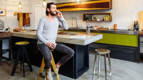 Person sitzt auf einem hohen Hocker an der Kücheninsel. Dahinter die moderne Küchenzeile.