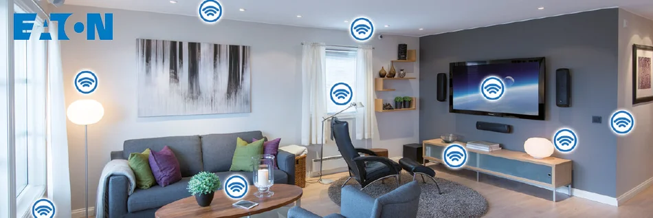 Das Bild zeigt einen gemütlichen Wohnraum mit einem Sofa. An einer Wand ist ein großer Bildschirm befestigt mit Lautsprechern rechts und links Darunter steht eine Konsole mit einer Lampe. Vor der Konsole steht ein Stuhl mit Armlehnen. Rechts und links vom Sofa stehen Stehleuchten. Im Bild sind Symbole für drahtlose Kommunikation bei den Lampen , dem Bildschirm und einem Wandschalter angebracht. 