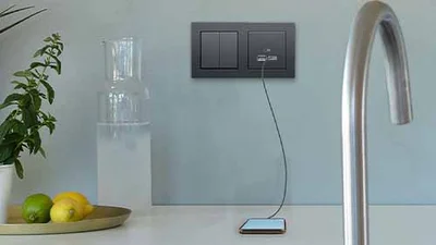 Vorne ein großer gebogener Wasserhahn in der Farbe Silber. Dahinter an der Wand montiert eine Kombination aus Steckdose und USB Ladegerät in der Farbe Schwarz.