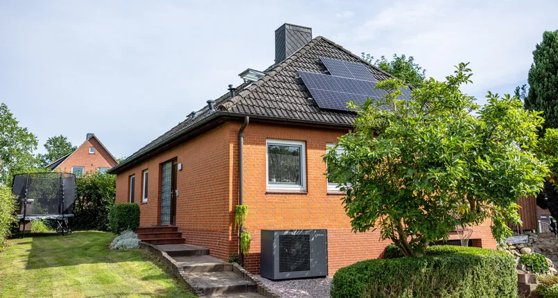 Das Bild zeigt ein Einfamilienhaus mit roter Klinkerfassade und dunkel gedecktem Satteldach. Auf dem Dach sind Solarmodule installiert. Im Vorgarten steht ein Baum und es sind gepflegte Hecken zu sehen. An der Hauswand ist eine Wärmepumpe der Marke Dimplex montiert. Im Hintergrund befindet sich ein weiteres Haus sowie ein Trampolin im Garten.
