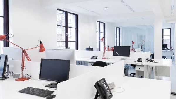 Das Bild zeigt einen großen Büroraum mit halbhohen, weißen Zwischenwänden zwischen einzelnen Schreibtischgruppen. Die Schreibtische sind weiß, Bildschirme, Tastaturen und Telefone auf den Schreibtischen sind schwarz. Die Schreibtischlampen sind rot. Die linke Wand des Raumes wird durch große Fenster mit schwarzen Fensterrahmen durchbrochen. Die Decke des Raumes ist weiß..