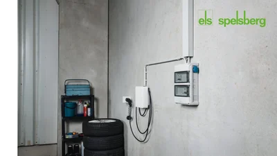 Das Bild zeigt eine an der Garagenwand montierte Wallbox mit zugehörigem Elektroverteiler. Im Vordergrund sind gestapelte Autoreifen und ein Regal mit Werkzeugen zu sehen, oben rechts befindet sich das Spelsberg-Logo.