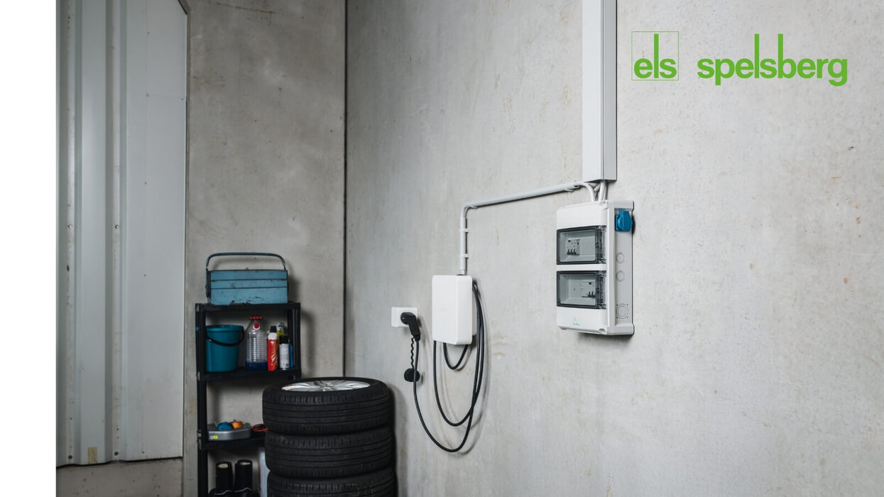 Das Bild zeigt eine an der Garagenwand montierte Wallbox mit zugehörigem Elektroverteiler. Im Vordergrund sind gestapelte Autoreifen und ein Regal mit Werkzeugen zu sehen, oben rechts befindet sich das Spelsberg-Logo.