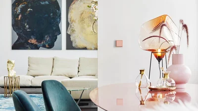Das Bild zeigt ein stilvoll eingerichtetes Wohnzimmer mit moderner Kunst an der Wand, einem beigen Sofa und dekorativen Accessoires auf einem Tisch. An der weißen Wand ist ein dezenter, quadratischer Schalter angebracht.