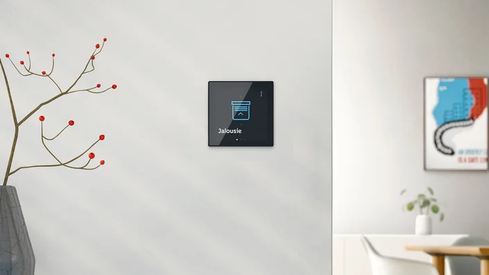 Das Bild zeigt einen modernen, hellen Innenraum mit einer weißen Wand, auf der ein Touchpanel mit der Anzeige "Jalousie" und einem entsprechenden Symbol befestigt ist. Links steht eine Vase mit dekorativen Zweigen, rechts sind ein Bild und ein Teil eines Tisches mit Stuhl zu sehen.