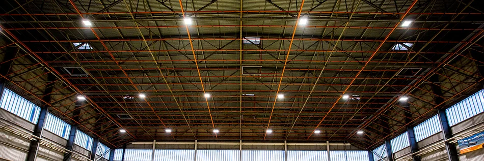 Das Bild zeigt eine große Halle mit Wellblechdach. Unter dem Dach ist ein Gestänge aus roten Metallverstrebungen befestigt. In regelmäßigen Abständen sind Hallenstrahler an Eckverbindungen der Verstrebungen angebracht.