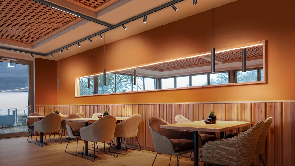 Das Bild zeigt den Innenraum eines Restaurants. Vor einer orangen Wand mit halbhoher Holzvertäfelung stehen drei Tische mit je vier Sesseln. Über Tischen hängt ein länglicher Spiegel. An der Decke sind Strahler an einem Schienensystem befestigt und setzen Lichteffekte an der Wand. Zusätzlich gibt es pro Tisch eine schmale, längliche Pendelleuchte, die ebenfalls am Schienensystem befestigt sind.