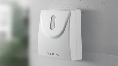 Das Bild zeigt einen weißen DEFENSOR Dämmerungsschalter von ESYLUX, der an einer weißen Betonwand befestigt ist. 
