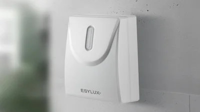 Das Bild zeigt einen weißen DEFENSOR Dämmerungsschalter von ESYLUX, der an einer weißen Betonwand befestigt ist. 