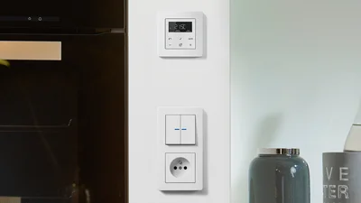 Fokus des Bildes liegt auf einem weißen JUNG-Schalter an der Wand. links im Bild ist ein Teil einer modernen Backofentür zu sehen und auf der rechten Seite Flaschen und Glasbehälter auf einer Ablage.