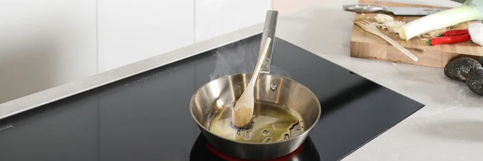 Das Bild zeigt eine Arbeitsfläche mit Ceranfeld. Auf dem Ceranfeld steht eine silberne Pfanne mit hohem Rand. Unter der Pfanne ist das Ceranfeld rot. In der Pfanne befindet sich Öl und ein Holzkochlöffel ist an den Pfannenrand gelehnt. Dampf steigt aus der Pfanne auf. Neben dem Kochfeld liegt ein Holzbrett mit Gemüse, einer Holzgabel und einem Messer.