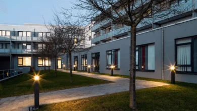 Das Bild zeigt den Innenhof eines modernen Gebäudeensembles bei Dämmerung. Im Hof verläuft ein Weg durch eine Rasenfläche mit vereinzelten Bäumen. Der Weg durch Pollerleuchten beleuchtet.