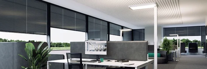 Das Bild zeigt ein großes, modernes Büro mit mehreren Arbeitsplätzen, großen Fenstern mit Jalousien, viel Tageslicht und einigen Pflanzen, wodurch eine freundliche Arbeitsumgebung entsteht.