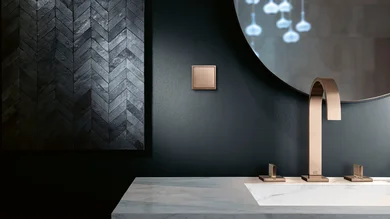 Das Bild zeigt ein modernes Badezimmer mit dunkler Wand, einem eckigen Wasserhahn und Armaturen in Bronze sowie einem passenden Lichtschalter. Die Einrichtung wirkt elegant und minimalistisch.