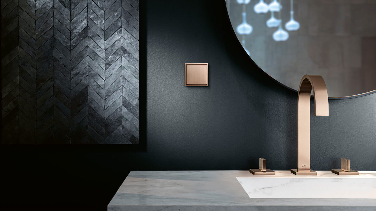 Das Bild zeigt ein modernes Badezimmer mit dunkler Wand, einem eckigen Wasserhahn und Armaturen in Bronze sowie einem passenden Lichtschalter. Die Einrichtung wirkt elegant und minimalistisch.