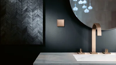 Das Bild zeigt ein modernes Badezimmer mit dunkler Wand, einem eckigen Wasserhahn und Armaturen in Bronze sowie einem passenden Lichtschalter. Die Einrichtung wirkt elegant und minimalistisch.