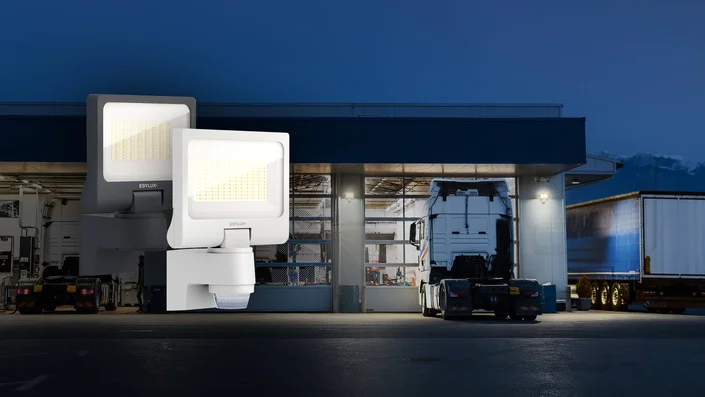 LED-Strahler mit integriertem Bewegungsmelder für die energieeffiziente Außenbeleuchtung von Gebäuden. Im Hintergrund ist ein beleuchteter Logistik- bzw. Werkstattbereich mit Lkw zu sehen.