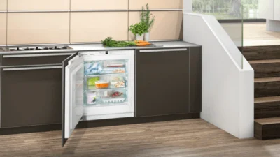 Ein geöffneter Unterbau Kühlschrank, der geöffnet ist und in einer braunen Küchenzeile integriert wurde. Daneben ein Treppenaufgang.