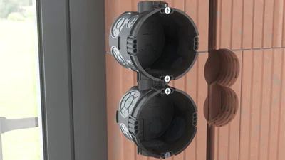 Zwei schwarze Unterputzdosen von OBO Bettermann, die sich vor einer noch unverputzten orangenen Wand befinden.