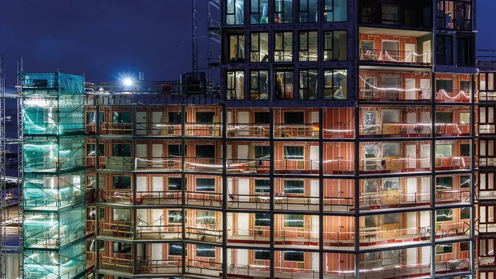 Das Bild zeigt die Baustelle eines mehrstöckigen Gebäudes bei Nacht. Die Fassade ist teilweise offen und beleuchtet, sodass die unfertigen Innenräume und die Gebäudestruktur sichtbar sind. Ein Teil des Gebäudes ist eingerüste