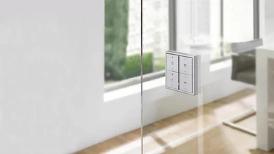 Das Bild zeigt ein modernes Smart-Home-Bedienpanel an einer Glaswand in einem hellen, minimalistisch eingerichteten Raum.