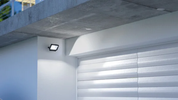 Das Bild zeigt die linke obere Ecke eines weißen Garagentores. Über der Garage gibt es einen Überstand. An der weißen Wand neben der Garage ist ein schwarzer OFL/AFL SUN LED Außenstrahler, der leuchtet. 