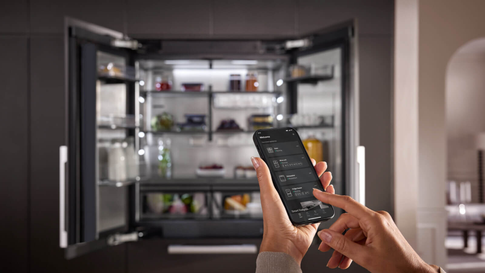 Eine Person bedient ein Smart-Home-fähiges Kühlgerät per App. Der offene Kühlschrank im Hintergrund ist modern gestaltet, mit LED-Beleuchtung und transparenten Fächern. Die App auf dem Smartphone ermöglicht die Steuerung von Funktionen wie Temperatur, Eismaschine oder Energieverbrauch.