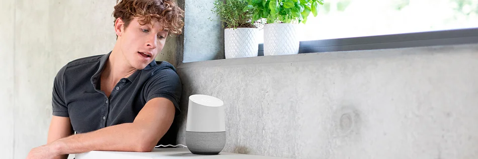 Das Bild zeigt eine Person, die sich zu einem Sprachassistenten auf einem Tisch wendet.