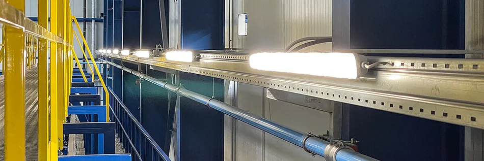Das Bild zeigt einen Industrieraum mit Rohren an der Decke und blau-gelben Metallgestängen. Vor den Fenstern sind eine Reihe von rechteckigen Feuchtraumleuchten an einer Metallschiene befestigt. 