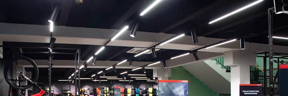 Das Bild zeigt die Decke eines Sportgeschäftes mit dicken Betonstützpfeilern. An der Decke sind an parallel verlaufenden Schienen längliche Leuchten und Strahlerleuchten im Wechsel installiert..