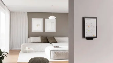 Das Bild zeigt ein modernes Wohnzimmer mit einem großen, hellen Sofa und minimalistischer Dekoration. An der Wand ist ein Smart-Home-Bedienpanel mit einer Benutzeroberfläche zur Steuerung verschiedener Funktionen angebracht.