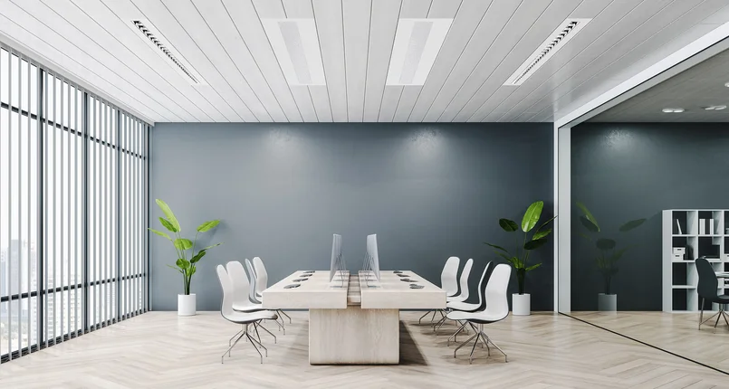 Ein modernes, helles Büro mit großem Tisch, mehreren Stühlen, Monitoren und großen Fensterfronten sowie Pflanzen an den Seiten.