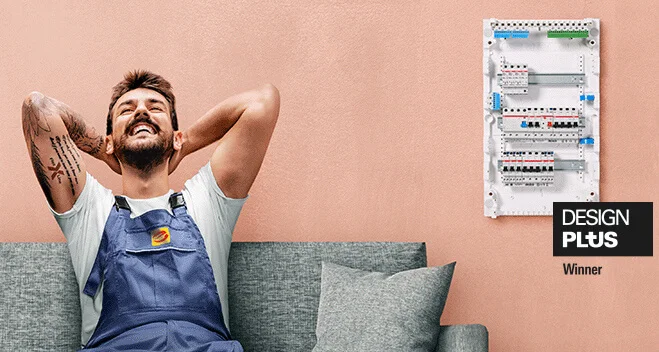 Das Bild zeigt eine Person, die entspannt auf einem grauen Sofa sitzt. Die Hände sind hinter dem Kopf verschränkt und der Blick nach oben gerichtet. Der rechte Arm der Person ist tätowiert. Die Person trägt ein weißes Shirt und eine graue Arbeitslatzhose. Auf der Hose ist das Zeichen für die Elektro-Innung aufgenäht. Rechts im Bild ist ein Verteilerkasten abgebildet. Über der rechten unteren Ecke des Kastens steht in einem schwarzen Rechteck Design Plus. Unter dem Rechteck steht Winner. Die Wand hinter der Person ist lachsfarben. 