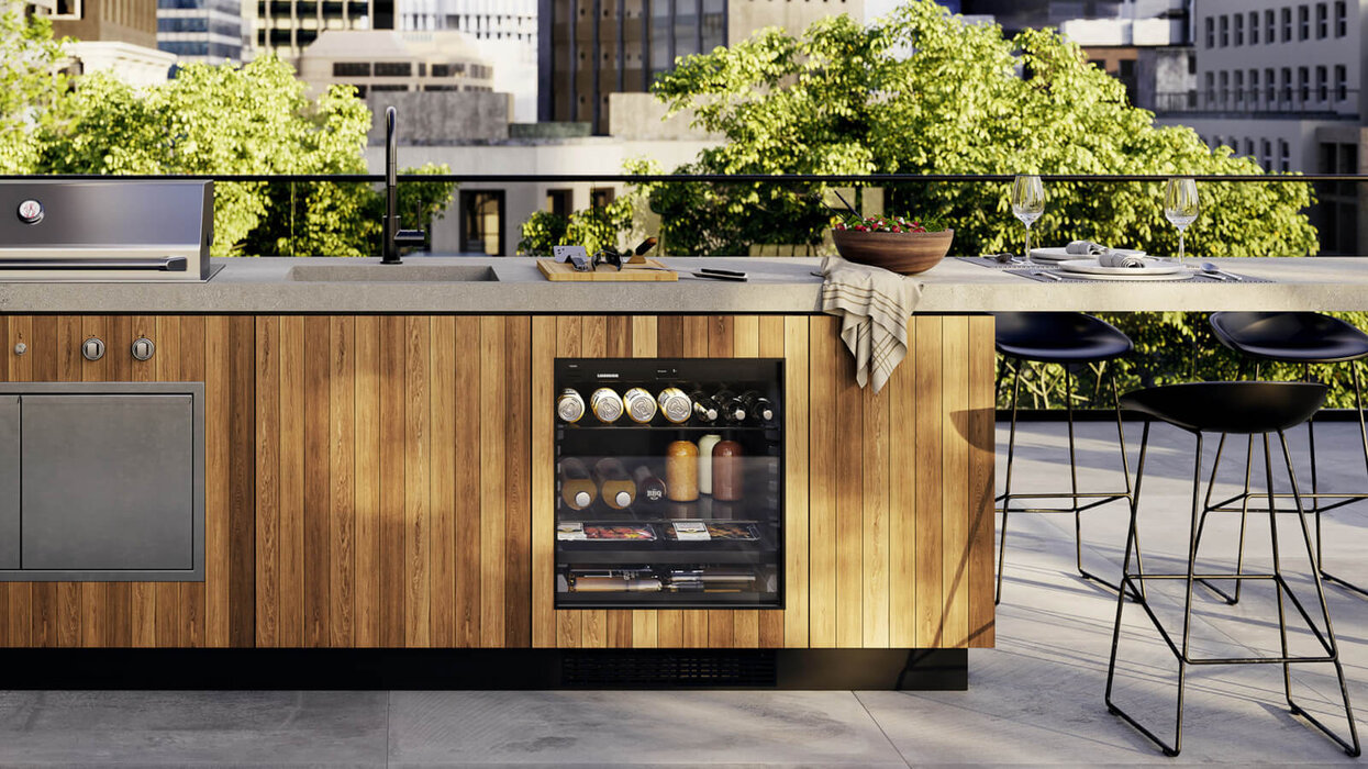 Außenküche mit Holzverkleidung, Arbeitsplatte, Grill, Spüle und integrierter Kühl-/Getränkeeinheit. Auf der Arbeitsfläche befinden sich Geschirr, Gläser und eine Schüssel, im Hintergrund sind Bäume und Gebäude zu sehen.