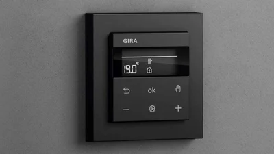 Das Bild zeigt einen schwarzen Gira System 3000 Raumtemperaturregler an einer dunkelgrauen Wand.