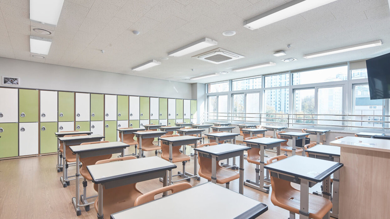 Das Bild zeigt ein helles Klassenzimmer mit Einzelpulten und Holzstühlen, grünen und weißen Spinden an der Rückwand, großen Fenstern auf der rechten Seite sowie mehreren rechteckigen Deckenleuchten.