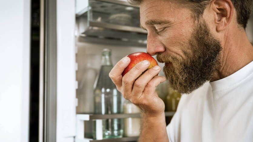 Eine Person hält einen Apfel vor die Nase und prüft dessen Frische vor einem geöffneten Kühlschrank.