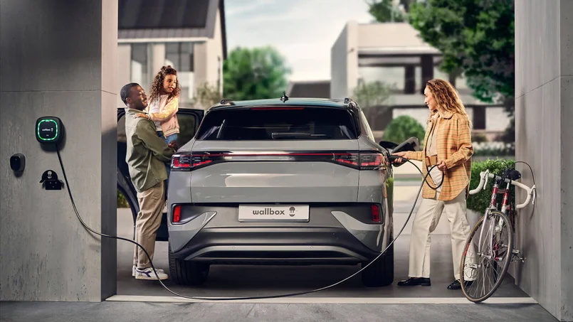 Das Bild zeigt ein Elektroauto, das in einer modernen Garage an einer Wallbox geladen wird. Eine Frau schließt das Ladekabel an, während ein Mann mit einem Kind am geöffneten Fahrzeug steht. Ein Fahrrad lehnt an der Wand.